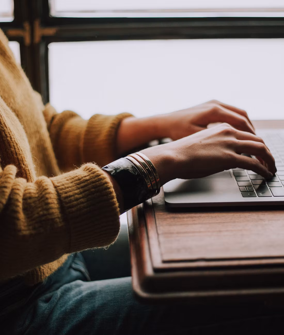 A woman in a mustard sweater typing on a laptop by a window.