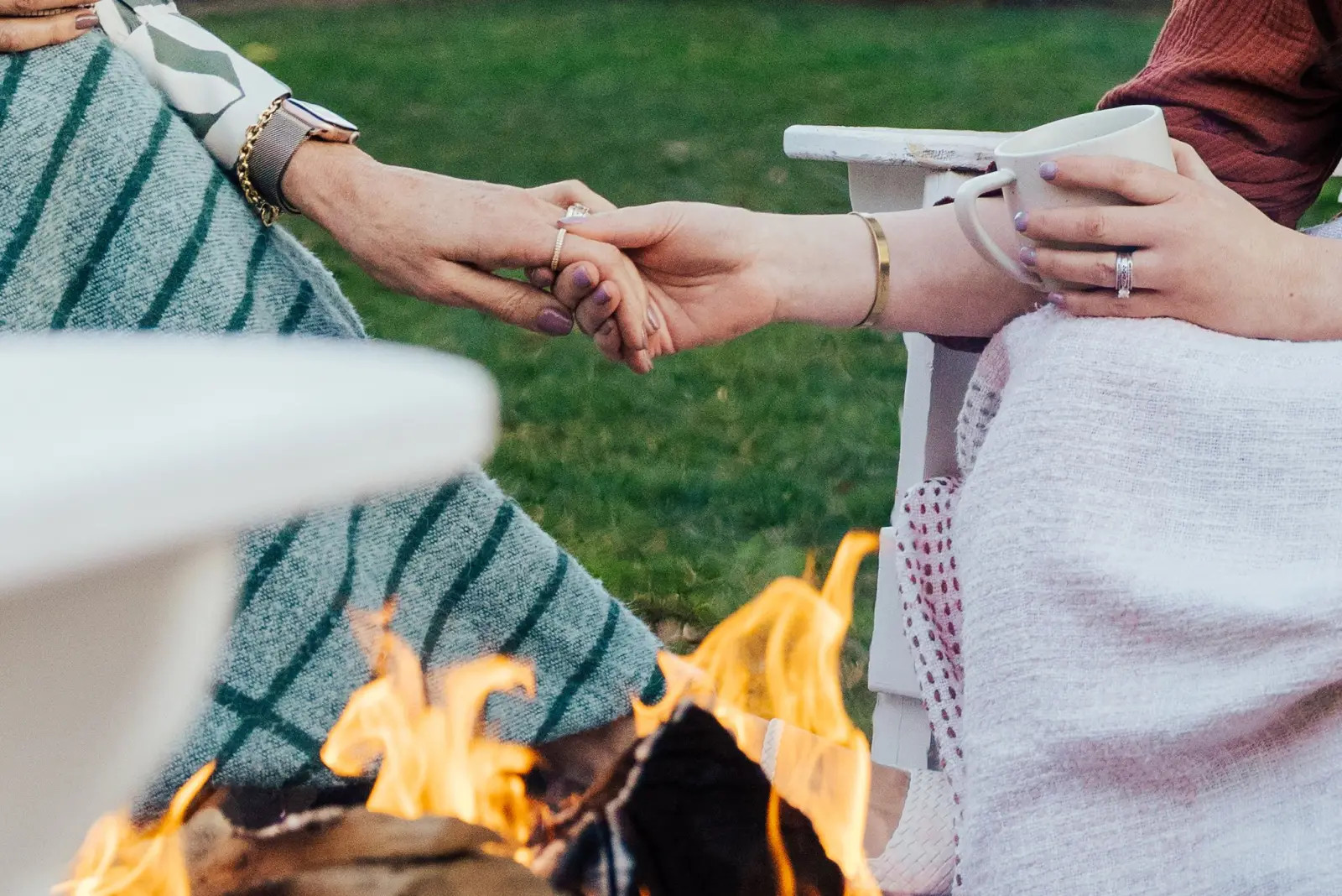 Two friends holding hands across a fire pit, wrapped in blankets, one holding a mug.