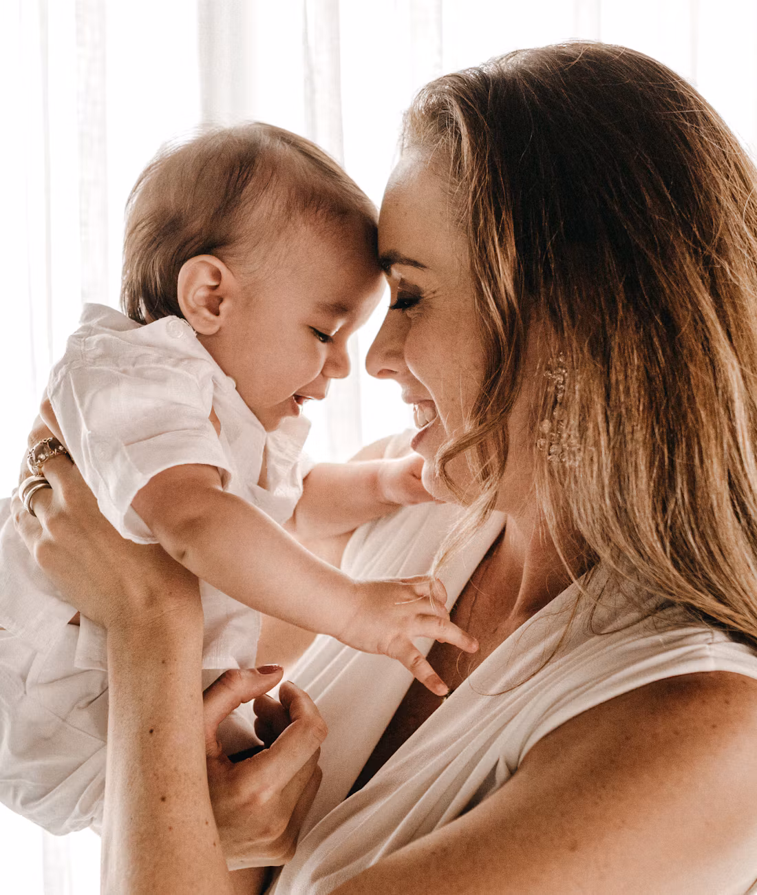 A smiling mother holding her baby up, their foreheads touching.
