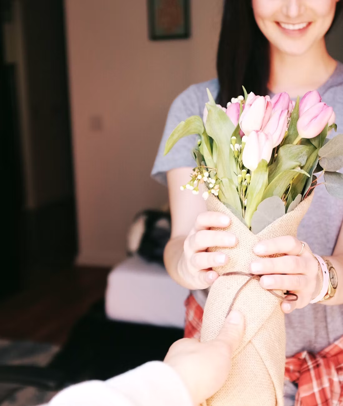 A friend handing a smiling woman a bouquet of pink tulips wrapped in burlap.