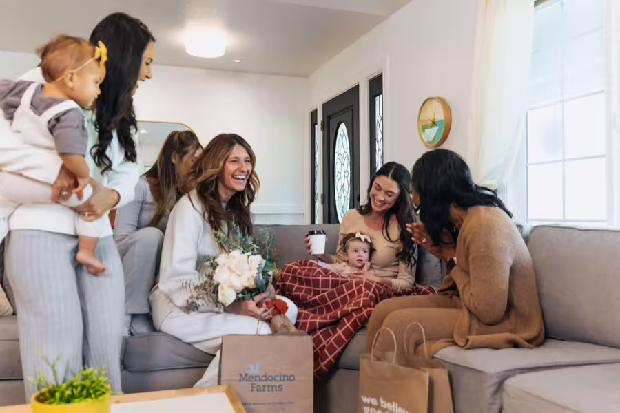 Friends visiting a new mom and her baby at home, with flowers and a box of food.